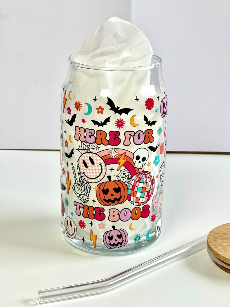 May include: A clear glass tumbler with a Halloween themed design. The tumbler features a colorful pattern of pumpkins, bats, skulls, and the text "Here for the boos".