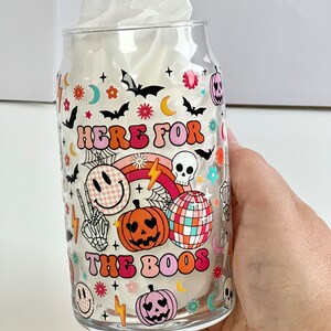 May include: A clear glass can with a Halloween themed design. The can has a pink, orange, and white design with the text "Here for the boos" and various Halloween icons.