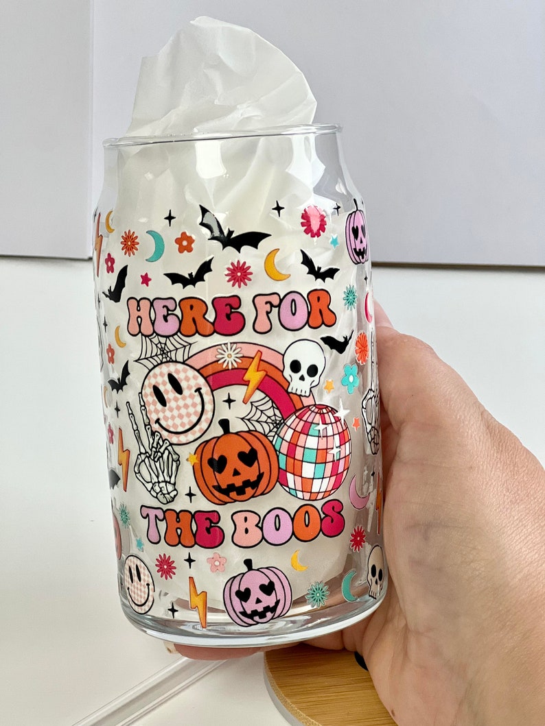 May include: A clear glass can with a Halloween themed design. The can has a pink, orange, and white design with the text "Here for the boos" and various Halloween icons.