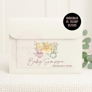 May include: Cream-coloured pregnancy notes organiser with a flap closure. The front features a watercolour illustration of baby clothes and the text "Baby Simpson Pregnancy Notes." A circular sticker reads "Available in other styles."