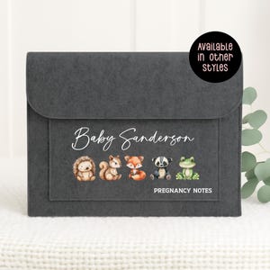 May include: A dark grey felt pregnancy notes organiser with a flap closure. The front pocket features watercolour illustrations of woodland animals and the text "Baby Sanderson" and "Pregnancy Notes."