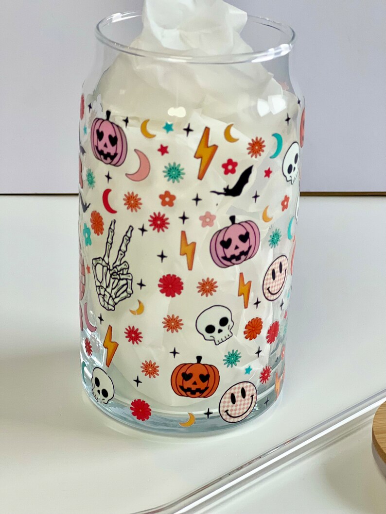 May include: A clear glass tumbler with a Halloween-themed design. The design features a variety of spooky and cute icons, including pumpkins, skulls, bats, stars, and lightning bolts. The tumbler is filled with a white tissue.