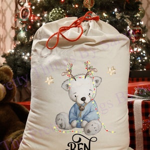 May include: An off-white Christmas sack with a red drawstring. It features a polar bear illustration wearing a blue sweater and reindeer antlers, with the name "BEN" printed in black script. The sack is set against a Christmas tree.