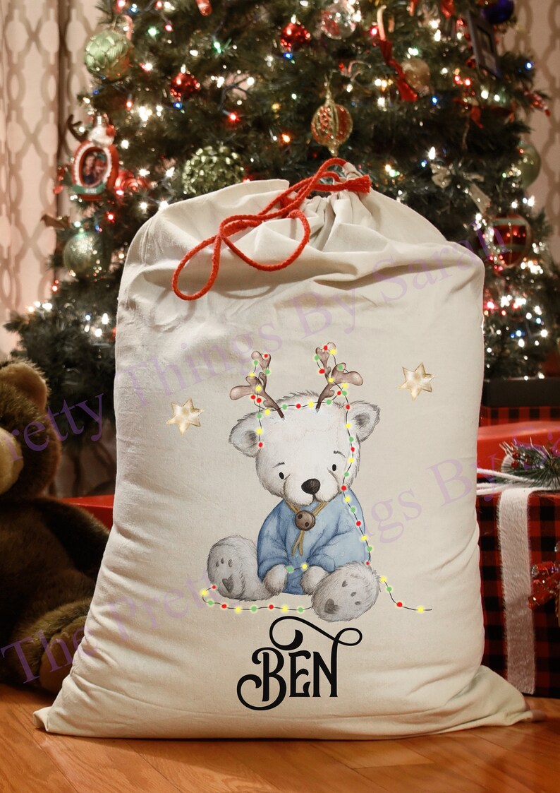 May include: An off-white Christmas sack with a red drawstring. It features a polar bear illustration wearing a blue sweater and reindeer antlers, with the name "BEN" printed in black script. The sack is set against a Christmas tree.
