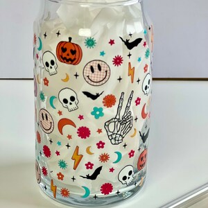 May include: A clear glass cup with a Halloween-themed design. The design features skulls, pumpkins, bats, smiley faces, stars, and crescent moons in a variety of colors, including red, orange, yellow, green, blue, and white.