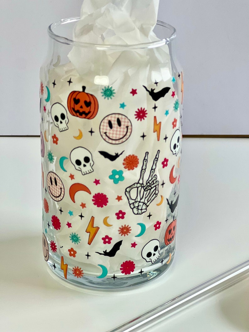 May include: A clear glass cup with a Halloween-themed design. The design features skulls, pumpkins, bats, smiley faces, stars, and crescent moons in a variety of colors, including red, orange, yellow, green, blue, and white.