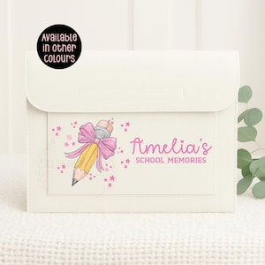 May include: Cream-coloured school memory book with a flap closure. The front pocket features a pencil illustration with a pink bow and the text "Amelia's SCHOOL MEMORIES". A circular sticker indicates the item is available in other colours.