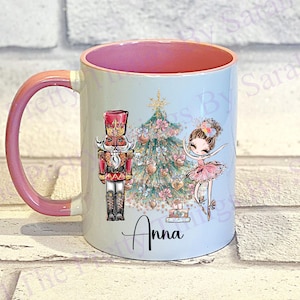 May include: A white ceramic mug with a pink interior and handle. The mug features a Christmas-themed design with a nutcracker, a decorated tree, and a ballerina. The name "Anna" is printed on the mug.