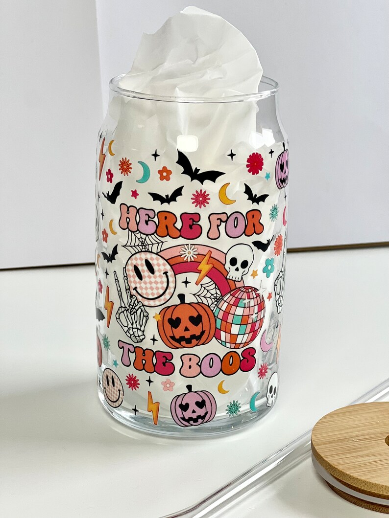 May include: A clear glass can with a Halloween themed design. The can has a white tissue inside and says "Here for the boos" with a rainbow, skulls, pumpkins, and smiley faces.