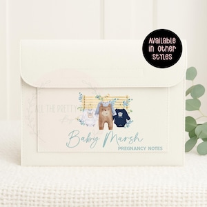 May include: Off-white pregnancy notes organiser with a flap closure. The front has a watercolour illustration of baby clothes and a bear. Text reads "Baby Marsh Pregnancy Notes". A black circle says "Available in other styles".