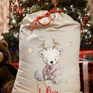 May include: An off-white Christmas gift sack with a red drawstring. It features a watercolour illustration of a white bear wearing a purple outfit and Christmas lights. The name "Olive" is printed in red.