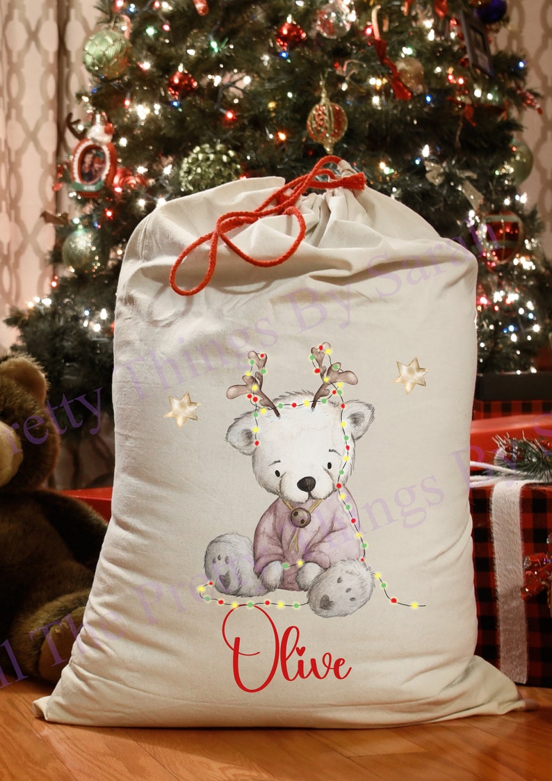 May include: An off-white Christmas gift sack with a red drawstring. It features a watercolour illustration of a white bear wearing a purple outfit and Christmas lights. The name "Olive" is printed in red.