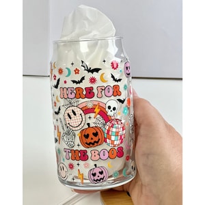 May include: A clear glass with a Halloween themed design. The glass has a pink, orange, and yellow design with a skull, pumpkins, and the text "Here for the boos".