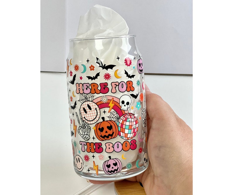 May include: A clear glass with a Halloween themed design. The glass has a pink, orange, and yellow design with a skull, pumpkins, and the text "Here for the boos".