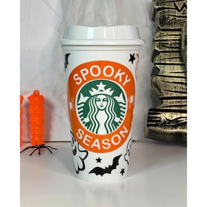 May include: White travel mug with a lid, featuring an orange band with the words "Spooky Season" around a green logo. The mug is decorated with black bats, spiders, ghosts, and stars, perfect for Halloween.
