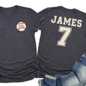 Könnte beinhalten: Zwei anthrazitfarbene T-Shirts. Eines hat eine Baseball-Grafik mit dem Namen "James" auf der Vorderseite. Das andere hat "JAMES" und die Nummer "7" auf der Rückseite. Ein Paar weiße Turnschuhe und blaue Jeans sind ebenfalls im Bild.