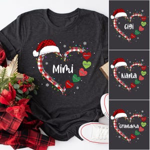 以下が含まれることがあります： 赤と白のチェック柄のハートのデザインが施されたダークグレーのTシャツ。ハートの上には赤いサンタの帽子が乗っており、クリスマスの電飾で囲まれています。ハートの中には「Mimi」という文字が書かれています。中には名前の書かれた小さなハートが5つあります。