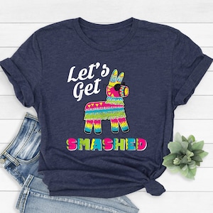 Puede incluir: Una camiseta azul marino con un gráfico blanco de una piñata con gafas de sol y el texto "Let's Get Smashed".