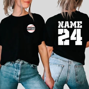 Könnte beinhalten: Schwarzes T-Shirt mit Baseball-Grafik und dem Text "TEAM NAME". Auf der Rückseite steht "NAME" über der Nummer "24" in Weiß. Das T-Shirt wird mit blauen Jeans getragen. Geeignet für Sportbegeisterte.