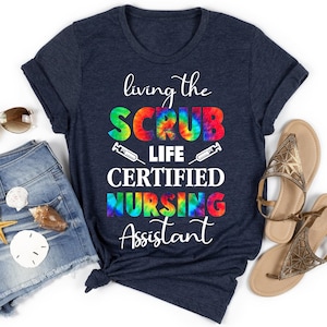 Op de afbeelding: Een marineblauw t-shirt met een tie-dye ontwerp met de tekst "living the SCRUB LIFE CERTIFIED NURSING Assistant".