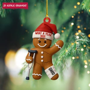 May include: A brown gingerbread man Christmas ornament with a red Santa hat and the text "WHAT A YEAR 2025". The gingerbread man has a leg in a cast with the name "DAVID" and is holding a crutch. The ornament is hanging from a gold string.