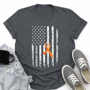 May include: A dark charcoal gray short-sleeve t-shirt featuring a distressed white American flag graphic. An orange awareness ribbon is centered over the stripes, and the text LEUKEMIA AWARENESS is printed vertically in white along the left side.