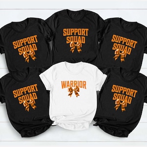 Può includere: Cinque magliette a maniche corte disposte in gruppo. Quattro magliette sono nere con il testo arancione SUPPORT SQUAD e un grafico a nastro arancione. Una maglietta bianca centrale presenta il testo arancione WARRIOR e un nastro arancione.