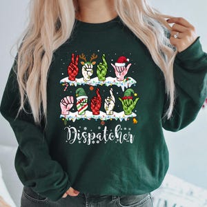 Könnte beinhalten: Dunkelgrünes Sweatshirt mit festlichem Design. Das Design zeigt Hände in Gebärdensprache, die weihnachtliche Formen bilden, mit dem Wort "Dispatcher" in weißer Schrift darunter. Das Design umfasst Rentiergeweihe, Weihnachtsmützen und Elfenhüte.