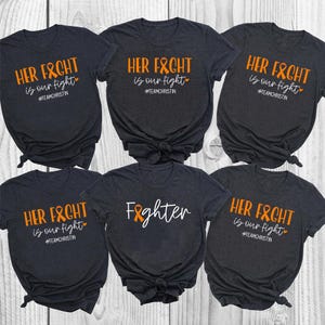 Può includere: Sei magliette grigio scuro. Cinque hanno la scritta "HER FIGHT is our fight" in arancione e bianco, con un cuore e l'hashtag #TEAMCHRISTIN. Una maglietta dice "Fighter" in bianco con un nastro arancione.