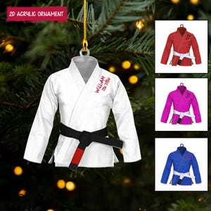 May include: A white 2D acrylic ornament shaped like a martial arts uniform, featuring a black belt and red accents. The text "WILLIAM JIU JITSU" is printed in red. Additional images show red, pink, and blue uniforms.
