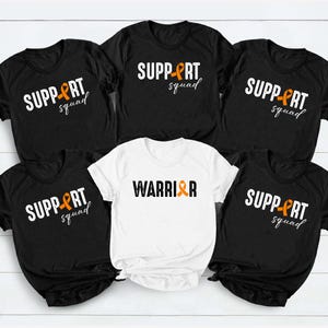 May include: Four black t-shirts and one white t-shirt. The black shirts feature the white text SUPPORT SQUAD with an orange ribbon replacing the letter O. The white shirt features the black text WARRIOR with an orange ribbon replacing the letter O.