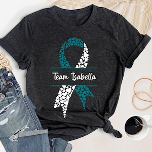May include: Dark gray t-shirt featuring a teal and white ribbon design made of hearts. The text "Team Isabella" is centered below the ribbon. The shirt is styled with rolled sleeves and a knot at the waist.