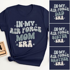 May include: Navy blue t-shirt with the text "IN MY AIR FORCE MOM ERA" in a retro font. Additional designs include "DAD ERA", "SISTER ERA", and "BROTHER ERA" on separate navy blue backgrounds.