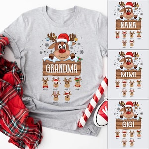 Pode incluir: Uma camiseta cinza com uma rena de desenho animado usando um chapéu de Papai Noel e segurando um cartaz que diz "Avó". Há seis rostos de rena menores abaixo do cartaz com espaços para nomes.