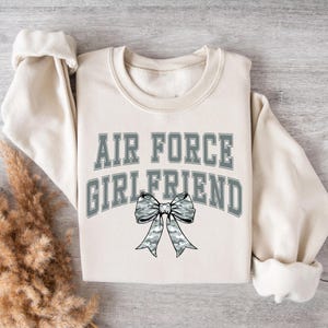 May include: A cream-colored sweatshirt with the text "AIR FORCE GIRLFRIEND" in gray letters. A gray and white camouflage bow is centered below the text.