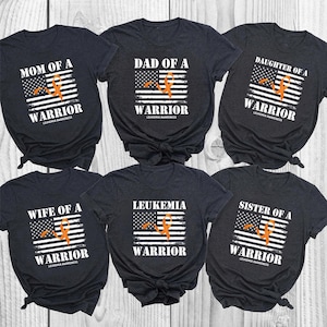 Può includere: Sei magliette grigio scuro con una grafica della bandiera americana bianca e un nastro arancione. Ogni maglietta riporta scritte come Mom, Dad, Daughter, Wife, Sister of a Warrior o Leukemia Warrior, con Leukemia Awareness stampato sotto.