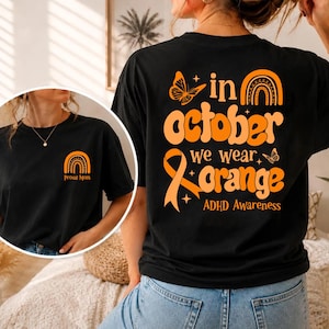 以下が含まれることがあります： オレンジ色のグラフィックが施された黒いTシャツ。背面には蝶、虹、啓発リボン、そして「in october we wear orange ADHD Awareness」という文字が描かれています。胸元には小さなオレンジ色の虹と「Proud Mom」という文字がプリントされています。