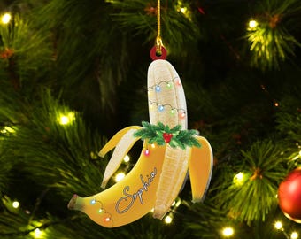 May include: A yellow banana Christmas ornament with a green holly sprig and colourful lights. The banana is split open and the word "Sophia" is written on the inside.