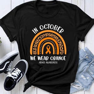 Könnte beinhalten: Ein schwarzes T-Shirt mit einer Grafik eines orangefarbenen Regenbogens mit Leopardenmuster und einer orangefarbenen Schleife in der Mitte. Der Text lautet IN OCTOBER WE WEAR ORANGE ADHD AWARENESS in weißen Großbuchstaben.