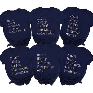 May include: Six navy blue t-shirts with phrases in colourful text. Phrases include "Most likely to laissez les bons temps rouler", "Most likely to find the king cake baby", and "Most likely to get lost on bourbon street".
