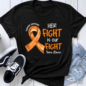 Könnte beinhalten: Ein schwarzes Kurzarm-T-Shirt mit einer orangefarbenen Schleife zur Leukämie-Aufklärung und dem weiß-orangen Text "LEUKEMIA AWARENESS HER FIGHT is our FIGHT Team Name" auf der Vorderseite. Das T-Shirt wird zusammen mit schwarz-weißen Turnschuhen gezeigt.