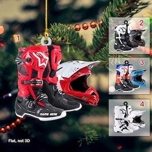 Puede incluir: Cuatro adornos navideños diferentes con botas y cascos de motocross. Los adornos son rojos, negros, blancos y azules. Las botas son todas negras con diferentes acentos de color. Los cascos son todos de diferentes colores y tienen una variedad de diseños. Los adornos son todos planos y no en 3D. El texto "NAME HERE" está impreso en las botas rojas y negras.