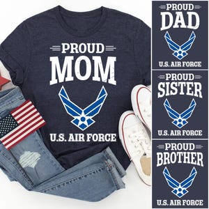 May include: A navy blue t-shirt with white text that reads "=PROUD= MOM" and a blue U.S. Air Force logo. The shirt is paired with blue jeans and white sneakers.