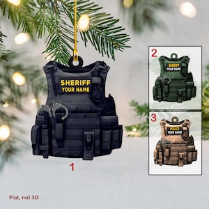 May include: A black, green, and tan Christmas ornament shaped like a tactical vest with the words "SHERIFF YOUR NAME" on the front. The ornament is flat and not 3D.
