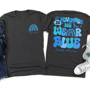 May include: Two dark gray t-shirts. One has a blue rainbow graphic and the text "Team Name". The other shirt has the text "November We Wear Blue Diabetes Awareness" in shades of blue, with a blue ribbon and butterfly graphic.