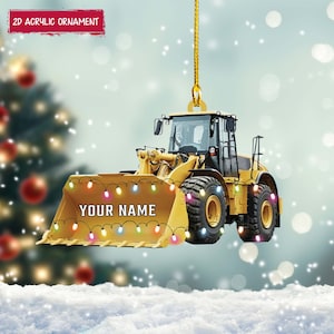 May include: A yellow front-end loader Christmas ornament with the text "YOUR NAME" on the bucket. The ornament is decorated with colorful lights and the text "2D ACRYLIC ORNAMENT" on a red banner. The ornament is hanging from a gold string.