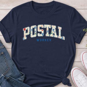 Puede incluir: Una camiseta azul marino de manga corta con la palabra POSTAL en letras grandes de estilo bloque rellenas con un patrón colorido de iconos y sellos relacionados con el correo. Debajo de la palabra POSTAL, la palabra WORKER está impresa en azul.
