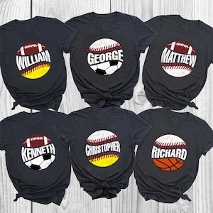 May include: Six dark grey t-shirts with sports-themed designs. Each shirt has a name printed on a ball graphic: William on a football, George on a baseball/soccer ball, Matthew on a football, Kenneth on a baseball/soccer ball, Christopher on a baseball/yellow ball, and Richard on a baseball/basketball.