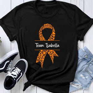 May include: A black short-sleeve t-shirt featuring a large awareness ribbon graphic composed of many small orange hearts. Across the center of the ribbon, the text "Team Isabella" is printed in a white script font between two horizontal lines.
