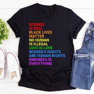 Könnte beinhalten: Schwarzes T-Shirt mit einer regenbogenfarbenen Botschaft, die lautet: "Wissenschaft ist real, Black Lives Matter, Kein Mensch ist illegal, Liebe ist Liebe, Frauenrechte sind Menschenrechte, Freundlichkeit ist alles."